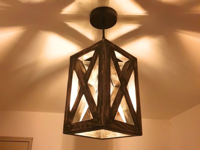 Plafonnier artisanal en pin massif finition noir mat vieilli, projetant des ombres géométriques chaleureuses au plafond.